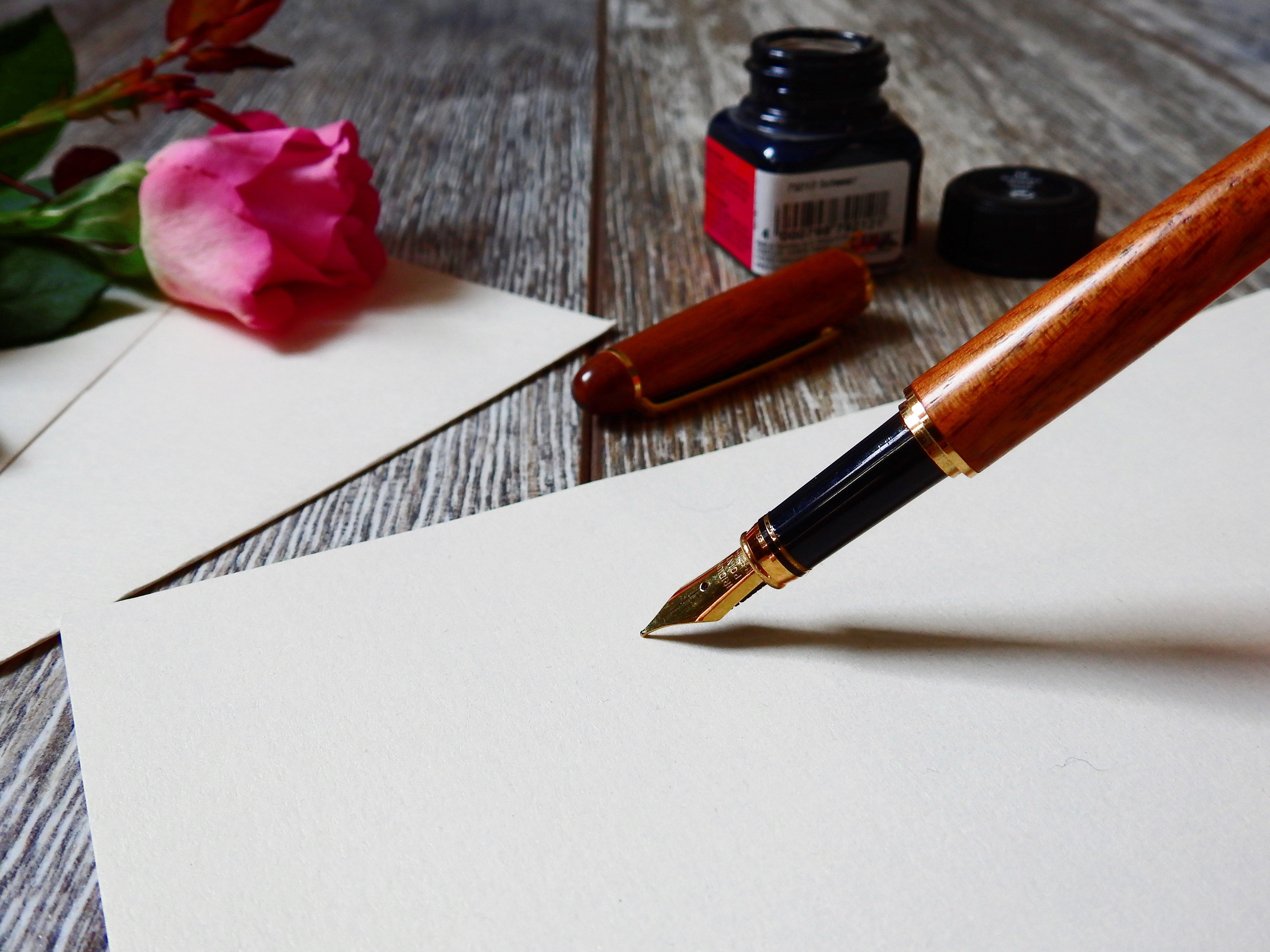 Wood fountain pen pressed against a blank piece of paper with cap, inkwell, and pink rose in the background